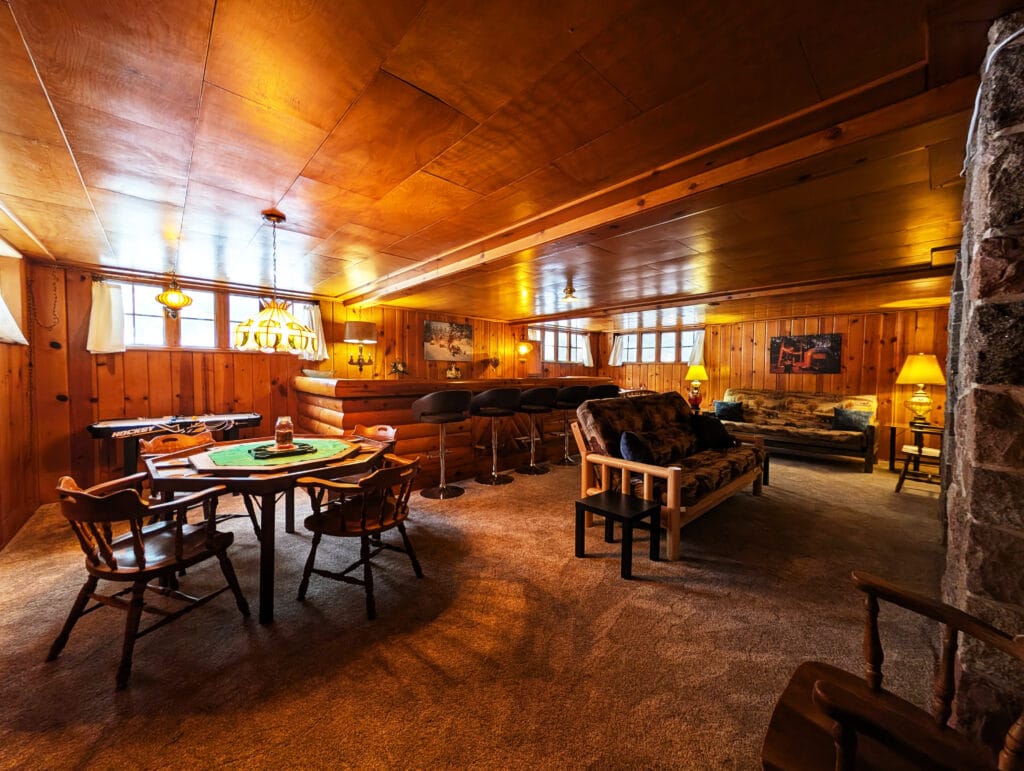Cozy rustic basement lounge with wood-paneled walls and ceiling, a pool table in the foreground, and warm lamps nearby.